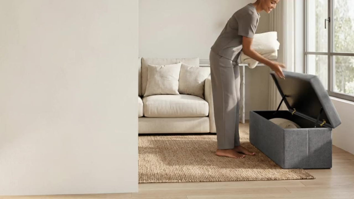 A woman stores a blanket inside a grey Fuho storage ottoman, closes the lid and sits on it